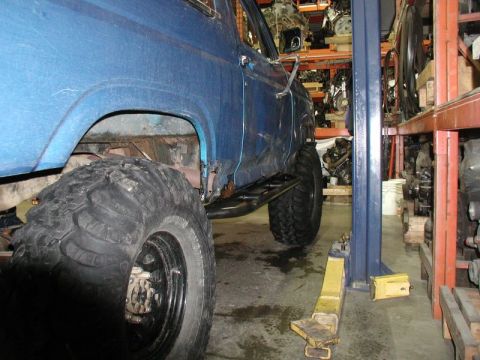 1985 Ford Bronco - The side view of a blue Ford Bronco is shown on a vehicle lift in a garage with shelves of car parts in the background. The vehicle has lar…