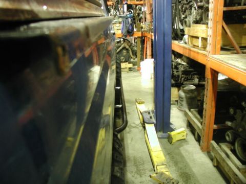 1985 Ford Bronco - A close-up, low-angle side view of a dark blue vehicle's fender and tire in a workshop setting. A yellow hydraulic jack is visible on the f…