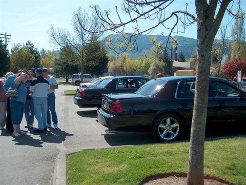 2001 Ford Crown Victoria - A group of people gather around two black sedans parked on a sunny day.