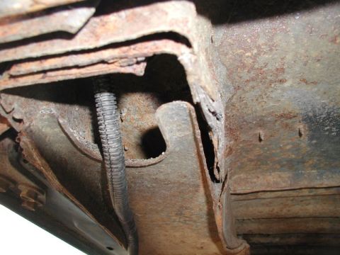 1985 Ford Bronco - The underside of a vehicle shows significant rust and corrosion on the frame and body panels. A black corrugated conduit runs along the rus…