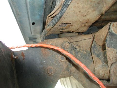 1985 Ford Bronco - Close-up view of the rusty undercarriage of a vehicle, showing corroded metal components and an orange wire running across the frame.