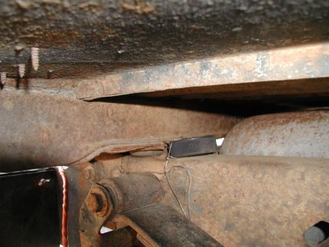 1985 Ford Bronco - A close-up view of the underside of a rusty vehicle frame, showing a small black rectangular object secured with wire to a metal bracket.