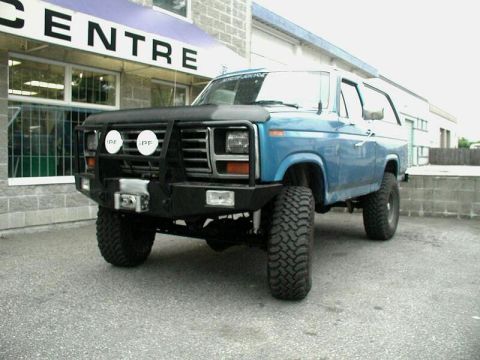 1985 Ford Bronco - Low 3/4 front view of a lifted blue Ford Bronco with a white top, black bug deflector, and a heavy black steel winch bumper with brush guar…