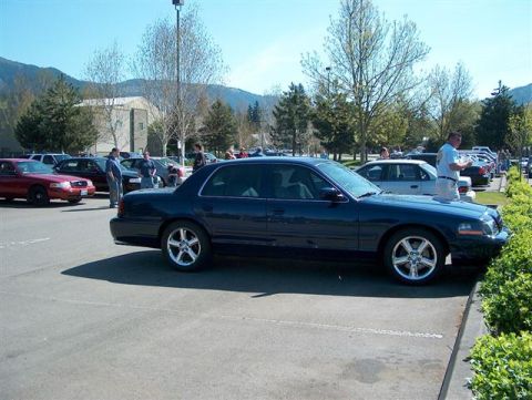 2001 Ford Crown Victoria - A dark blue 2001 Ford Crown Victoria is parked in a lot with several other vehicles and people milling about.