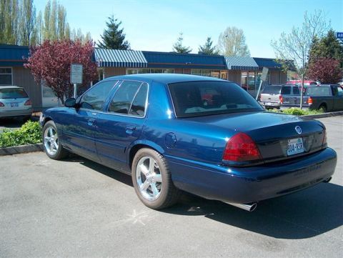 2001 Ford Crown Victoria - A blue 2001 Ford Crown Victoria is parked in a lot with other vehicles and a building in the background.