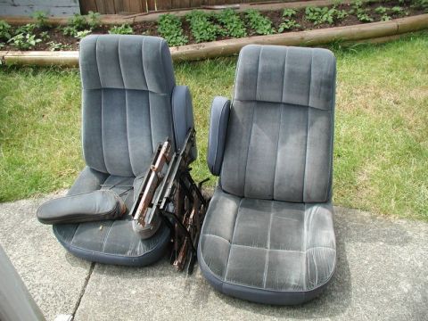 1985 Ford Bronco - Two dark gray velour car seats with armrests are displayed outdoors on a concrete surface next to a grassy area and a wooden fence.