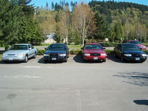 2001 Ford Crown Victoria - Four sedans are parked in a lot, including a silver 2001 Ford Crown Victoria.