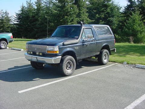 1985 Ford Bronco - A dark blue Ford Bronco is parked in a paved lot with trees in the background. The vehicle is viewed from the front 3/4 angle, showing its…