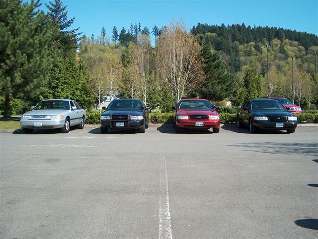 Four police sedans parked in a lot. The silver one on the left looks older than the three dark ones on the right