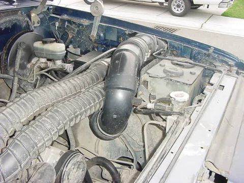 1985 Ford Bronco - The engine bay of a dark blue vehicle is shown, featuring a large black intake tube, ribbed hoses, and a brake fluid reservoir.