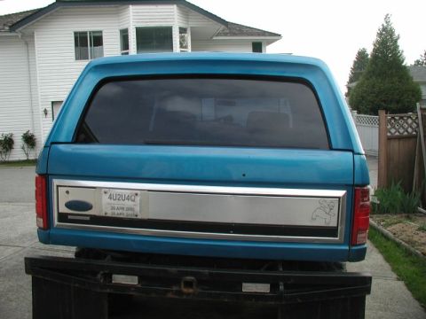 1985 Ford Bronco - Straight-on rear view of a teal-blue Ford Bronco with a matching hardtop and tinted rear glass, showing the brushed-aluminum tailgate trim…