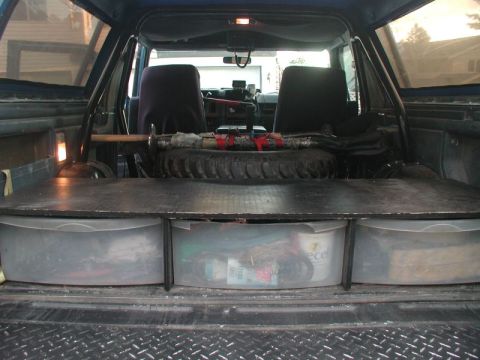 1985 Ford Bronco - The interior rear of a blue Ford Bronco shows a custom storage unit with three clear plastic bins beneath a flat wooden surface. A spare ti…