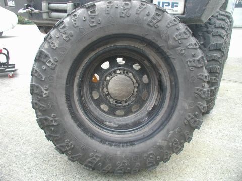 1985 Ford Bronco - A close-up, low-angle view of a large, aggressive off-road tire mounted on a black rim. The tire has prominent tread blocks and text that r…