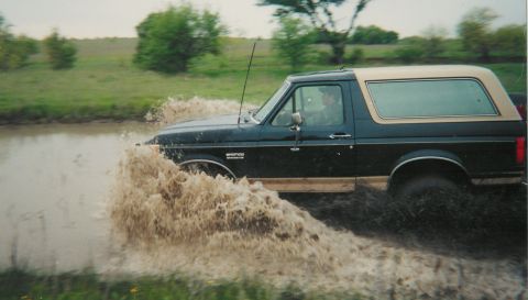 1991 Ford Bronco - Scanned print of a dark blue Ford Bronco with a tan hardtop running left-to-right through a muddy water hole, blasting a thick sheet of bro…