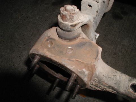 1985 Ford Bronco - A close-up view of a rusty, cast metal vehicle suspension component, likely a steering knuckle or control arm, with several threaded bolts…