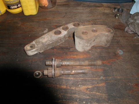 1985 Ford Bronco - Two rusty metal brackets and two threaded bolts with nuts and washers are laid out on a wooden workbench. A yellow bottle with a black cap…
