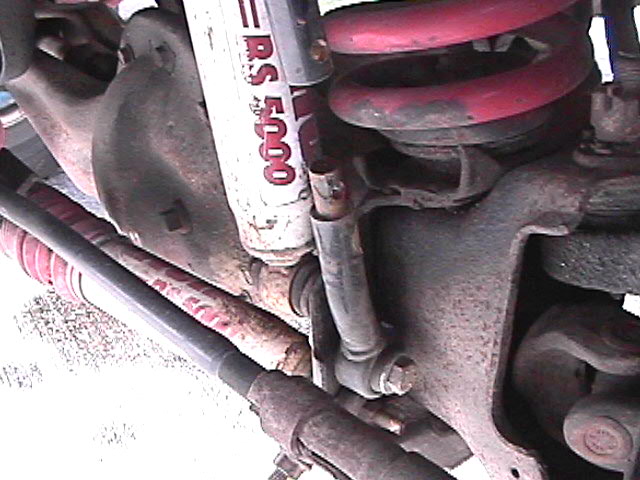 Vehicle suspension with red coil spring, Rancho shock, and tie rod on rusty differential.