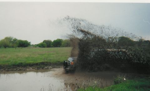 1991 Ford Bronco - Scanned print of a dark 4x4 charging through a mud hole at the edge of a green pasture, hurling a huge arc of brown mud and water over the…