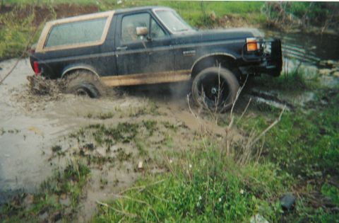 1991 Ford Bronco - Scanned print of a dark blue Ford Bronco with tan two-tone trim pulling out of a shallow muddy creek crossing, rear tire slinging mud and w…