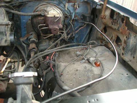 1985 Ford Bronco - The engine bay of a blue vehicle, showing the brake master cylinder, various hoses, wiring, and a rusted firewall.