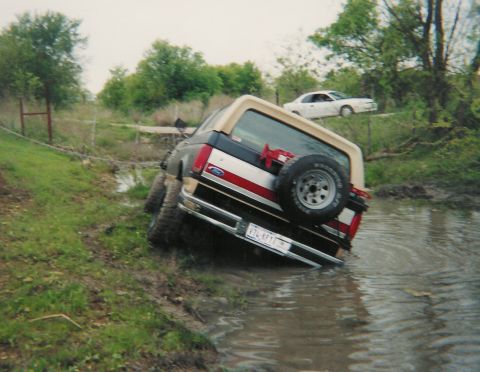 1991 Ford Bronco - Rear 3/4 view of a two-tone dark blue and tan Ford Bronco with a tan hardtop tipped over onto its passenger side into a muddy creek, driver…