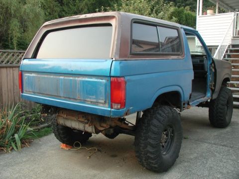 1985 Ford Bronco - The rear driver's side of a lifted, two-door SUV with large off-road tires. The vehicle is blue with a brown roof and appears to be undergo…