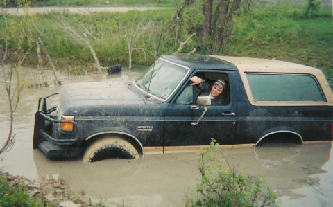 1991 Ford Bronco - Dark blue Ford Bronco with a tan hardtop sits buried to its rocker panels in a muddy creek crossing, water lapping over the tires and front…