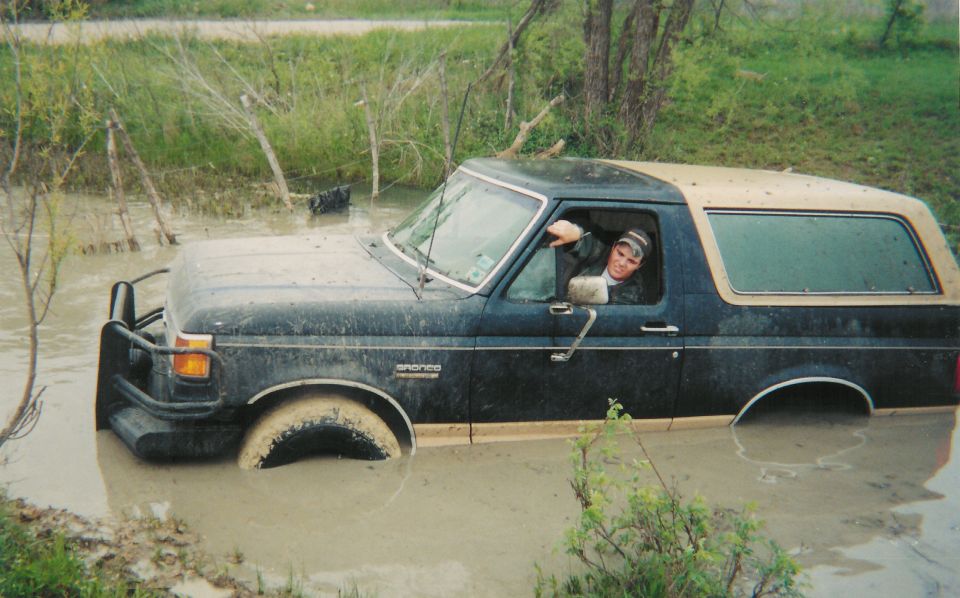 Dark blue Ford Bronco with tan top stuck axle-deep in a muddy creek, driver waving from the window