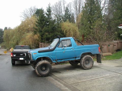 1985 Ford Bronco - A lifted, bright blue 1980s Ford Bronco with large off-road tires and a snorkel air intake is parked on a concrete driveway next to a black…