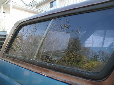 1985 Ford Bronco - A close-up side view of the rear window of a blue and brown Ford Bronco. The window is tinted and reflects trees and a house.