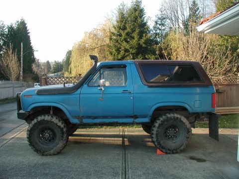 1985 Ford Bronco - The side profile of a lifted blue 1980s Ford Bronco is shown parked on a concrete driveway. The vehicle features large off-road tires, a sn…
