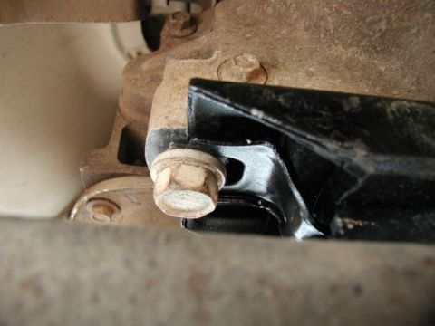 1985 Ford Bronco - A close-up, low-angle shot shows a bolt securing a black metal bracket to a rusty, tan-colored vehicle component. The bolt head is hexagona…
