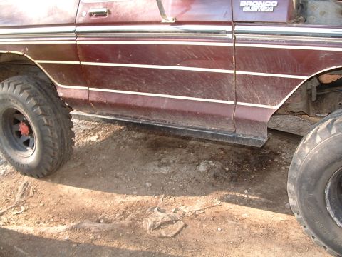 1979 Ford Bronco - Close side view of a maroon Ford Bronco's lower body on dirt, showing the "BRONCO CUSTOM" badge, pinstriped side molding and a flat black s…
