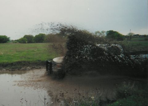 1991 Ford Bronco - A dark-colored SUV with a front brush/grille guard plows through a large muddy water pit, throwing a massive spray of mud and water into th…