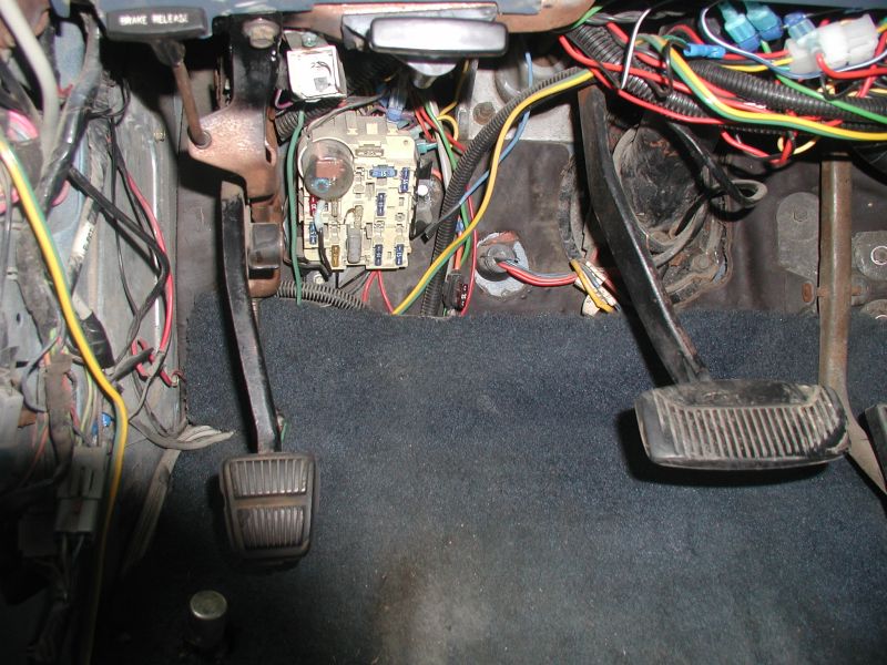 Interior view of vehicle footwell with pedals, fuse box, and wiring.