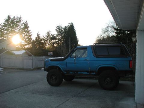 1985 Ford Bronco - A blue Ford Bronco with off-road tires and a snorkel is parked on a driveway next to a house. The sun is setting behind trees in the backgr…