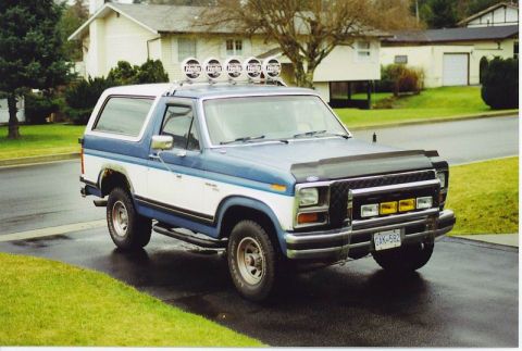1985 Ford Bronco - A blue and white two-door Ford Bronco is parked on a wet asphalt driveway in front of a residential home. The Bronco features a roof-mounte…