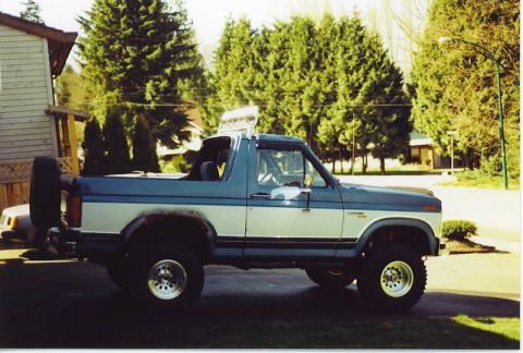 1985 Ford Bronco - The side view of a lifted, two-tone blue and white Ford Bronco is parked on a paved surface in front of a house and trees. A spare tire is…