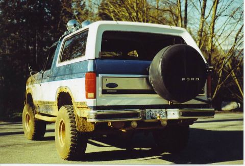 1985 Ford Bronco - The rear passenger side of a lifted, two-tone blue and white Ford Bronco is shown parked on a paved surface with trees in the background. I…