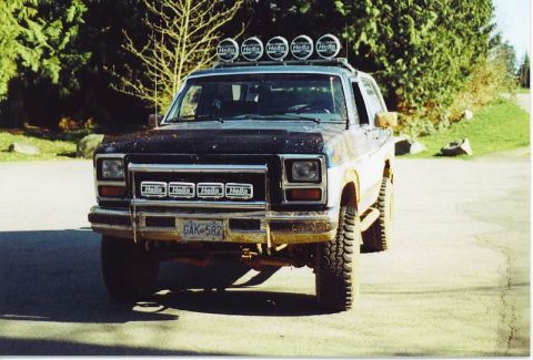 1985 Ford Bronco - A muddy, dark blue and white 1985 Ford Bronco with five Hella lights mounted on the roof rack and four Hella lights on the front grille. Th…