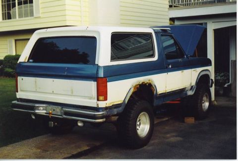 1985 Ford Bronco - The rear driver's side of a two-tone blue and white Ford Bronco is shown parked in a driveway. The vehicle has significant rust on the rear…