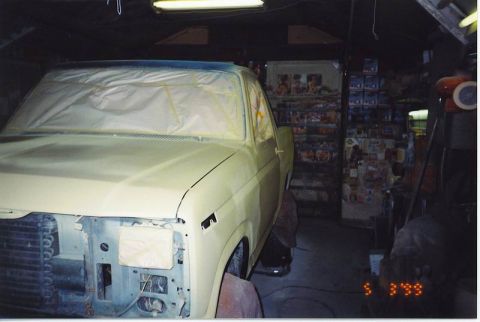1985 Ford Bronco - A light yellow pickup truck is parked in a garage, with its front end covered in masking paper and tape, likely for painting. Shelves stock…
