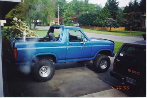 1985 Ford Bronco - A lifted, bright blue 1980s Ford Bronco is parked next to a black GMC pickup truck, with smoke billowing from the Bronco's exhaust pipe. Th…