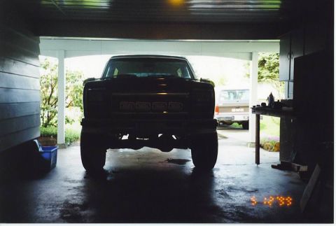 1985 Ford Bronco - The front of a dark-colored, lifted pickup truck is parked inside a garage. Another pickup truck, possibly a GMC, is visible in the backgro…