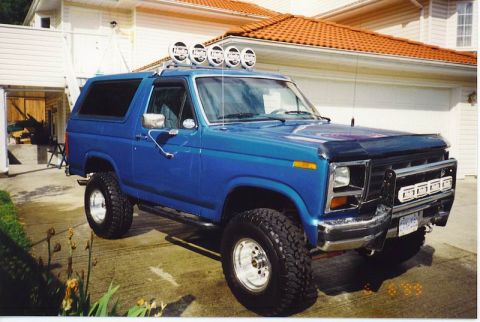1985 Ford Bronco - A lifted blue 1980s Ford Bronco is parked on a concrete driveway in front of a house with a red tile roof. The vehicle features a roof-moun…
