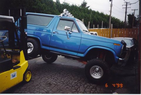 1985 Ford Bronco - A lifted, bright blue 1980s Ford Bronco is shown from the side, with its front end angled upwards. A yellow forklift is positioned to the l…