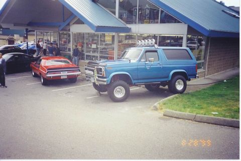 1985 Ford Bronco - A lifted blue Ford Bronco with off-road lights on the roof rack is parked in a lot in front of a building with large windows. A red muscle…