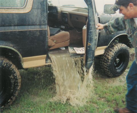1991 Ford Bronco - Muddy water pours out of the open driver's door of a dark blue two-tone Ford Bronco after a deep water crossing, flooding onto the grass wh…