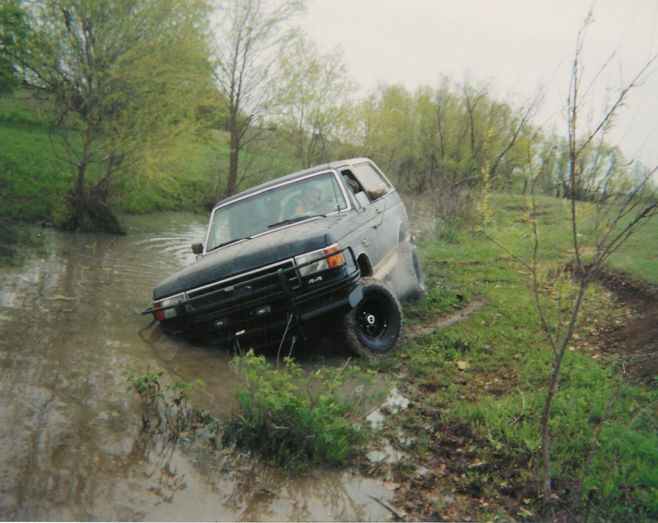 Blue and silver Ford Bronco climbing a muddy bank out of a flooded creek