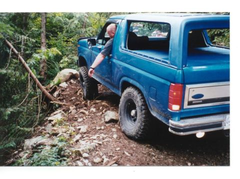 1985 Ford Bronco - A blue 1980s Ford Bronco is navigating a rocky, wooded trail with its driver's arm resting on the open door. The vehicle is positioned on a…